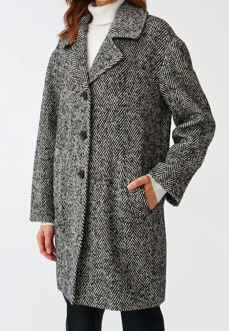 Kvinde iført en sort-hvid herringbone mønstret frakke med stor krave og sorte knapper, hånd i lommen, over hvid turtleneck og sorte bukser.