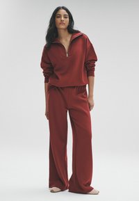 Bordová mikina na zips s rolovanými rukávmi a voľnými nohavicami v rovnakom odtieni. Mäkká látka s minimálnym dizajnom. Nosí sa s obuvou svetlých tónov.