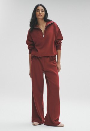 Maronfarvet zip-up sweatshirt med rulle ærmer og matchende løstsiddende bukser. Blødt stof med minimal design. Bæres med lysfarvet fodtøj.