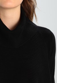 Schwarzer gerippter Strickpullover mit hohem Kragen und strukturiertem Muster. Die Nahaufnahme hebt den Stoff und die Designdetails hervor.