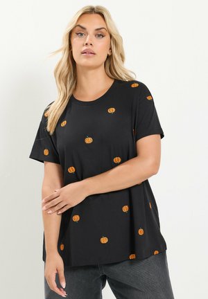 Vrouw draagt een zwart shirt met korte mouwen met kleine oranje pompoenpatronen, gecombineerd met donkergrijze broek, staande voor een effen achtergrond.