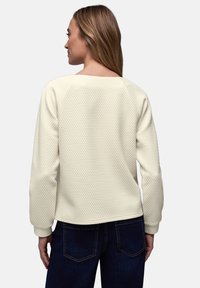Femme aux cheveux longs portant un pull texturé crème clair et un jean bleu foncé, vue de dos sur un fond uni.