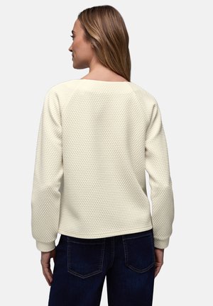 Femme aux cheveux longs portant un pull texturé crème clair et un jean bleu foncé, vue de dos sur un fond uni.