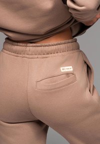 Braune Sweatpants mit elastischem Bund, Seitentasche und einem glatten, strukturierten Stoff für zusätzlichen Komfort. Sichtbares Markenetikett auf der Rückseite.