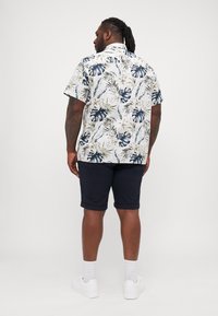 Camisa de manga corta con fondo blanco decorada con hojas verdes y marino, acompañada de pantalones cortos marinos y zapatillas blancas.