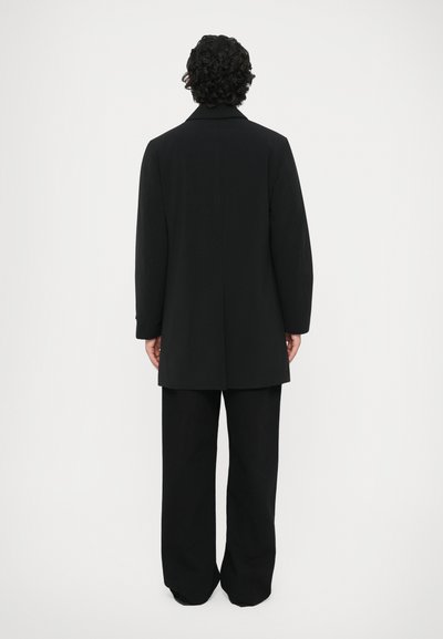 Zwarte overjas met een rechte snit, met notch-lapels en lange mouwen. Gecombineerd met zwarte broek met een brede pijp. Glad textuur, minimalistisch ontwerp.