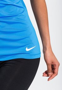 Niebieski sportowy top na ramiączkach marki Nike, wykonany z lekkiego, gładkiego materiału, z białym logo na dolnym brzegu. Czarne legginsy dopełniają look.