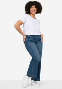 Camiseta blanca de manga corta, jeans de tiro alto y pernera acampanada en azul, y zapatillas blancas. Los jeans tienen un dobladillo con un sutil detalle deshilachado.