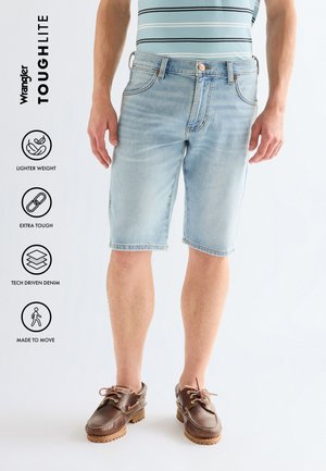 Man draagt lichtblauwe denim shorts en bruine leren bootschoenen, staand tegen een effen lichte achtergrond.