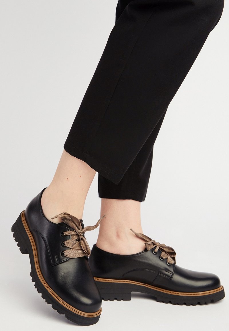 Personne portant des chaussures en cuir noir à lacets avec des semelles épaisses marron et des lacets beiges, associées à un pantalon noir court sur un fond blanc.