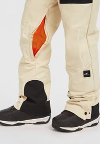 Pantalones de nieve beige con ventilaciones laterales con cremallera que revelan un forro de malla naranja, con acentos negros y parches de rodilla negros reforzados. Botas negras con suela texturizada.