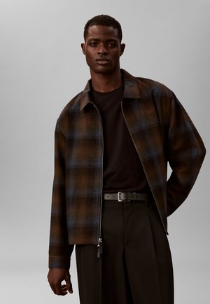 Chaqueta de cuadros marrón y azul con cierre de cremallera, combinada con una camisa y pantalones negros. Cuenta con cuello y costuras visibles.