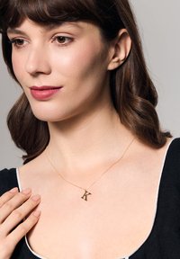 Złoty naszyjnik z zawieszką w kształcie litery "K", cienki łańcuszek, gładka tekstura, noszony na czarnej bluzce z białym obrzeżem, widoczny odcień skóry.