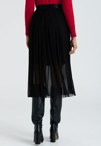 Svart plisserad midikjol med halvgennomsiktig tyg, elastiskt midjeband, kombinerad med figurnära svarta knähöga stövlar. Slät textur.