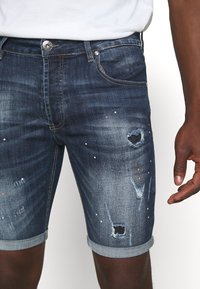 Bermudas de mezclilla en azul oscuro con detalles desgastados, múltiples pequeños agujeros, salpicaduras de pintura blanca y naranja, y dobladillos enrollados.