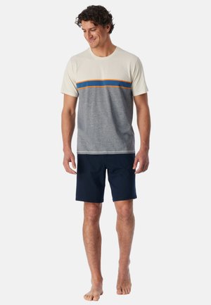 Man staat op blote voeten, draagt een beige-grijs T-shirt met korte mouwen met blauwe en oranje strepen en marineblauwe korte broek, kijkt naar beneden met een lichte glimlach.