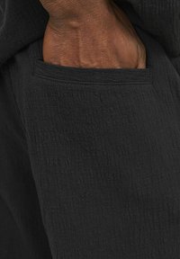 Tissu noir texturé avec une apparence douce et froissée. Gros plan sur une main insérée dans la poche, mettant en valeur les détails de couture.