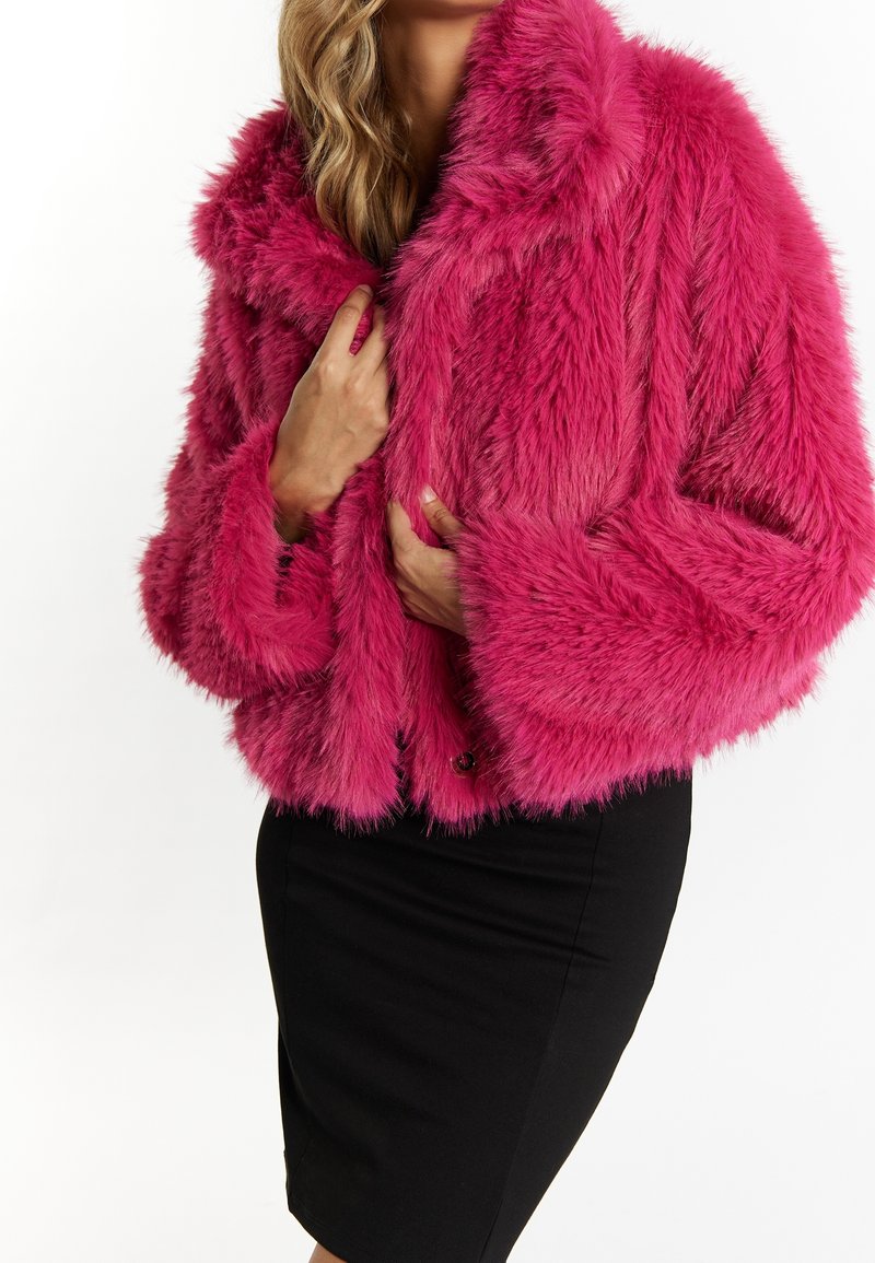 Femme portant une veste en fausse fourrure rose vif sur une jupe crayon noire, tenant le col de la veste avec les deux mains.