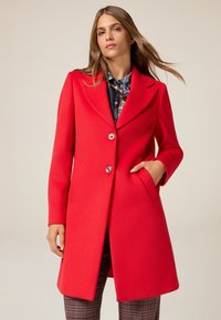 Abrigo de lana rojo con cuello, diseño de un solo botón, dos botones, bolsillos laterales y una textura suave, llevado sobre una camisa estampada.