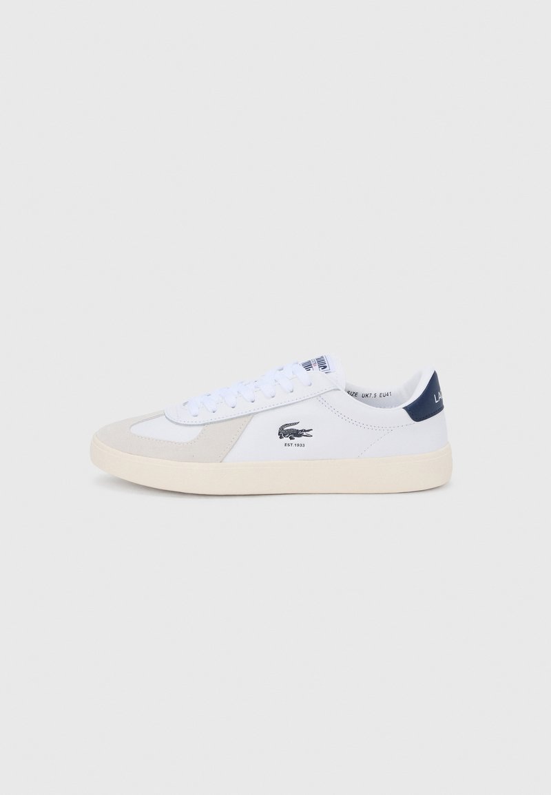 Hvid lavtop-sneaker med beige ruskindsdetaljer, navy hælflag, snørebånd foran og sort krokodillelogo på siden med "EST. 1933".