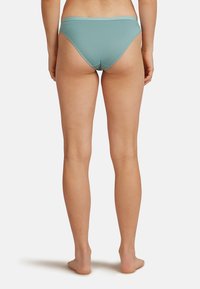 Person som står barfota med ryggen mot kameran, klädd i ljusblå baddräkt i bikini-stil mot en enkel vit bakgrund.