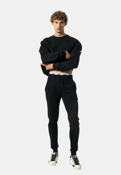 Junge Person mit lockigem Haar, die einen schwarzen Pullover, schwarze Jogginghosen, einen weißen, gebrandeten Bund und weiße Sneakers mit schwarzen Schnürsenkeln trägt und aufrecht steht.