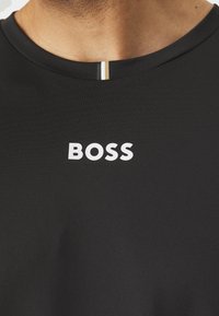 Juodas sportinis marškinėliai su tekstūruotu paviršiumi, su baltu "BOSS" logotipu ir juostele šalia apykaklės.