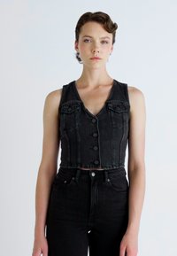 Gilet en denim noir court avec un col en V, cinq boutons de fermeture, et deux poches poitrine, associé à un jean en denim noir taille haute.