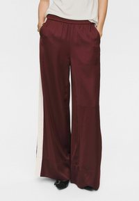 Pantaloni larghi in satin bordeaux con vita elastica e tasche laterali, caratterizzati da un pannello laterale di colore chiaro a contrasto.