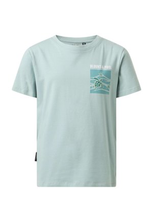 Garçon portant un t-shirt graphique bleu clair, un jean bleu et des baskets blanches, tenant une veste noire sur son épaule, debout sur un fond uni.