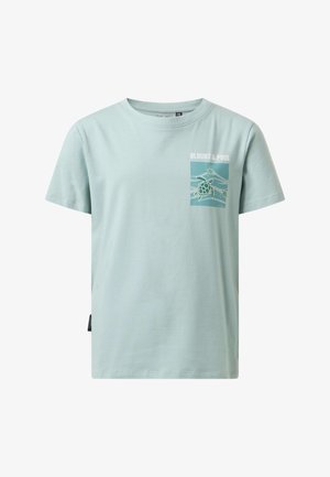 Dreng iført lyseblå t-shirt med grafik, blå jeans og hvide sneakers, der holder en sort jakke over sin skulder og står på en ensfarvet baggrund.