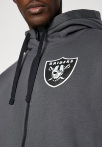 Szara bluza z kapturem na zamek błyskawiczny z czarnymi sznurkami, z dużym logo Raiders na klatce piersiowej. Wykonana z miękkiego materiału o gładkiej fakturze.