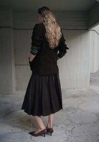 Chaqueta de ante marrón sobre una falda oscura plisada; tacones puntiagudos marrones. Textura suave, diseño en capas y silueta fluida.