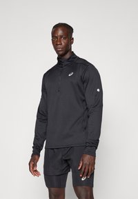 Svart atletisk långärmad tröja med dragkedja i halsen, med en logotyp, strukturerad tyg och figurnära design, ihop med svarta shorts.