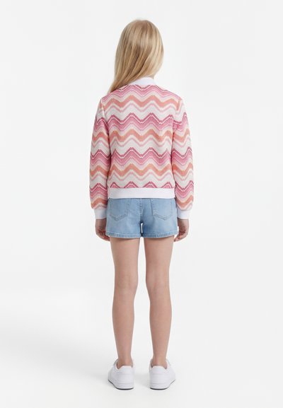 Enfant aux longs cheveux blonds portant un pull chevron rose et orange, un short en jean clair et des baskets blanches, debout de dos.