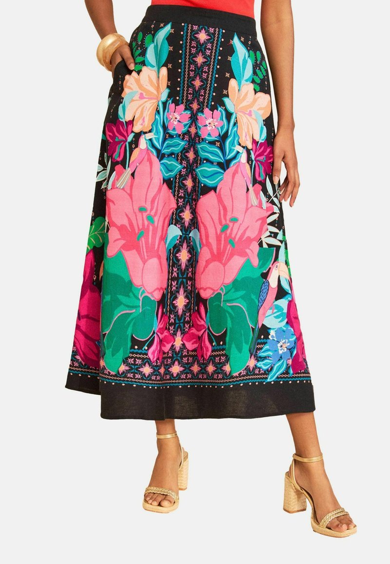 Black midi skirt with bright pink, green, blue floral and bird print, worn with beige high-heeled sandals and a red top.
