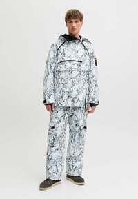 Jeune homme portant une veste à capuche et un pantalon à motifs marbrés noir et blanc avec des bottes marron, se tenant contre un fond clair uni.