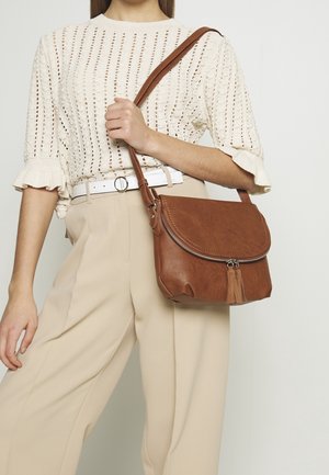Kvinde iført beige bukser og hvid striktrøje holder brun lædertaske med tværrem og nydetalje i form af kvast på lynlåsen på skulderen.