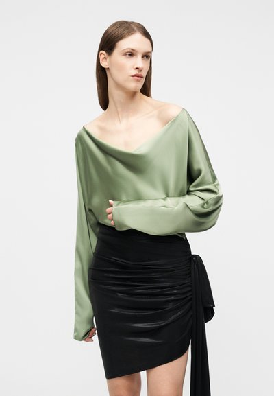 Γυναίκα με ίσια καστανή τρίχα που φοράει μια χαλαρή πράσινη σατέν μπλούζα έξω από τους ώμους και μια μαύρη δερμάτινη φούστα με ρέλια και δεσίματα στο πλάι.