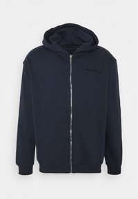 Marinblå zip-up hoodie i mjukt tyg, med en framficka, ribbade manschetter och en minimal logotyp på bröstet. Slät textur.