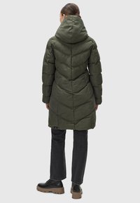 Zeleni puffer plašč s kapuco, ki ima kvadratno zasnovo, srednje dolg kroj in rahlo sijaj. Nošen z črnimi kavbojkami in masivnimi črnimi čevlji.