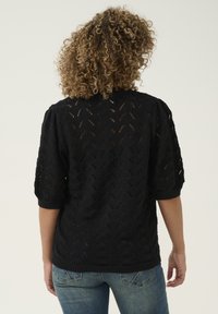 Pull noir en tricot avec un motif texturé de feuilles, manches courtes bouffantes et ourlet côtelé. Porté avec un jean bleu, affichant une coupe décontractée.