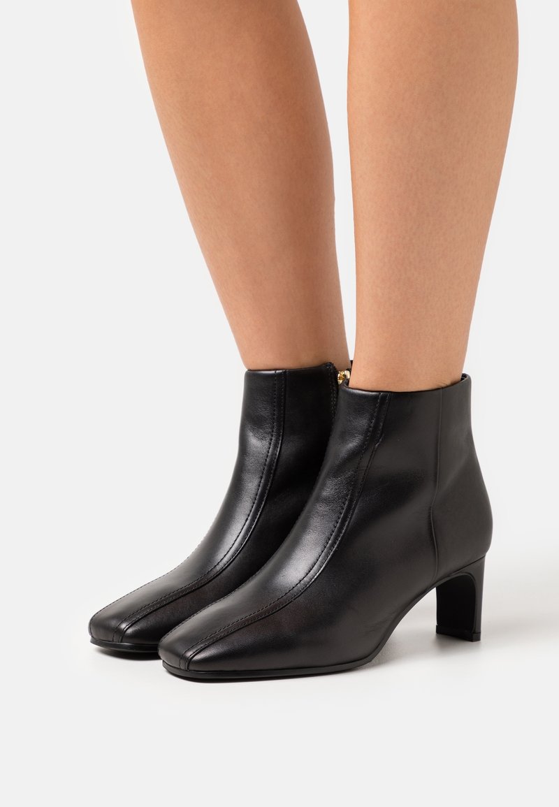 Botas de cuero negras hasta el tobillo con punta afilada, tacón alto delgado y cremallera lateral. Textura suave con detalles de costura.