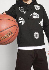 Henkilö mustassa hupparissa, jossa on NBA-joukkueen logoja, pitää koriin palloa kameraa kohti, rajattuna kaulasta keskivartaloon yksinkertaista taustaa vasten.