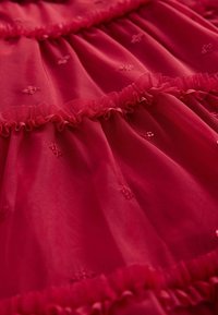 Tessuto rosso trasparente con piccoli punti ricamati e dettagli di arricciature orizzontali, che creano un effetto stratificato e testurizzato.