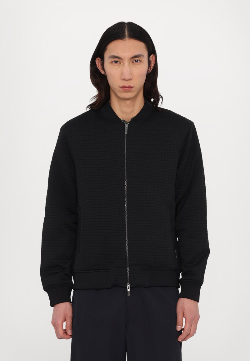 Giubbotto bomber trapuntato nero con zip anteriore, polsini e orlo a costine. Presenta un colletto rialzato e tasche sui lati. Design pulito e minimale.