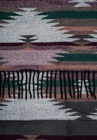 Couverture tissée présentant un motif géométrique dans des tons de vert, rose, gris et blanc, avec des franges noires le long du bord inférieur.