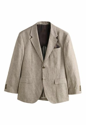 Homme aux cheveux courts, debout face à un mur blanc texturé, portant un costume marron clair à fines rayures, près d'escaliers en pierre à l'extérieur.