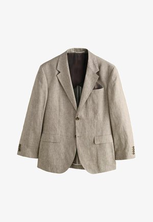 Mand med kort hår står vendt mod en hvid tekstureret væg iført en lys brun jakkesæt med nålestriber, nær stenetrin udendørs.