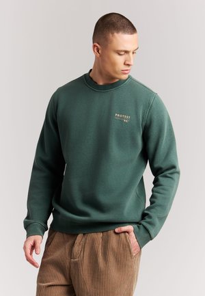 Man in een groene crewneck trui met "Protest"-logo en bruine corduroy broek, kijkt naar links met één hand in zijn zak.